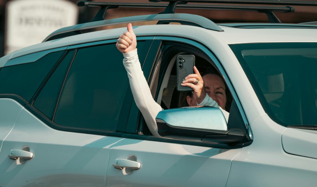 A woman gives a thumbs up while taking a photo with a smartphone from inside a car.