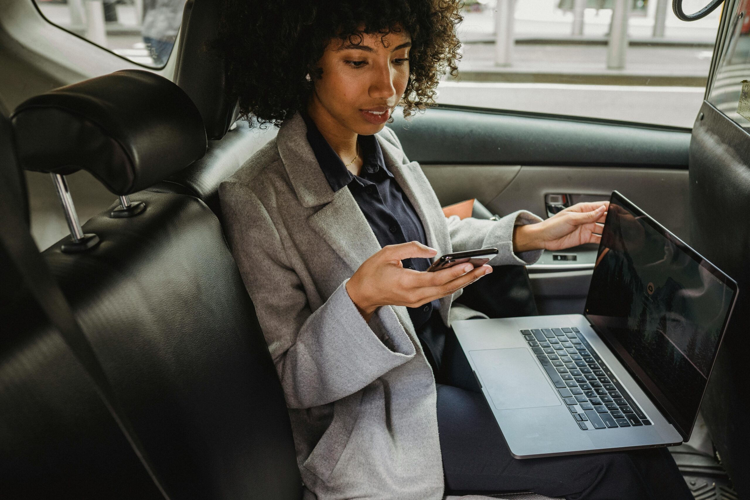 Crop African American female in formal clothes sitting with laptop on knees and browsing mobile phone in comfortable automobile